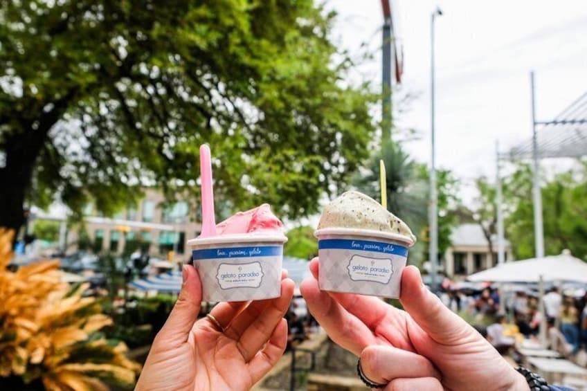 Two hands holding gelato cups with straws, one labeled 'gelato porocchio' and the other 'gelato paradiso,' in an outdoor setting with trees and a market in the background. 2