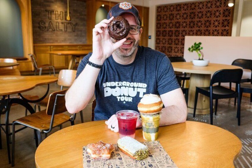 A person sitting at a table with a donut, a drink, and a sandwich. 1