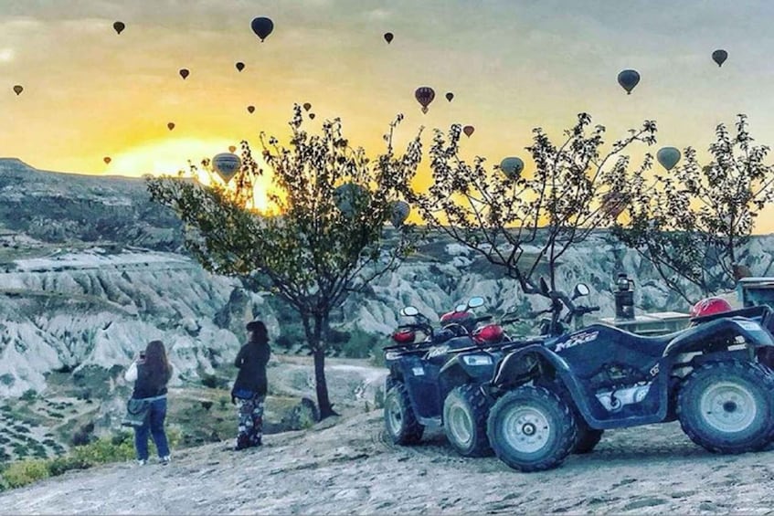 A quad bike parked on a rocky terrain with a sunset in the background and hot air balloons in the sky. 1
