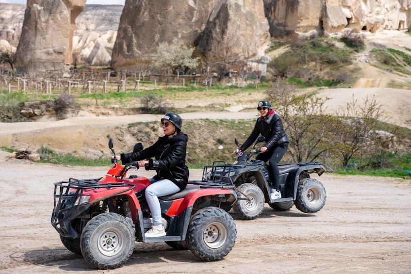 Two people riding ATVs on a dirt path with rock formations in the background. 3