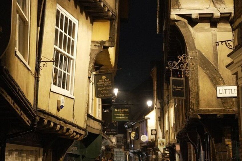 Una calle estrecha iluminada de noche, con letreros luminosos, incluyendo uno de una chocolatería. 2