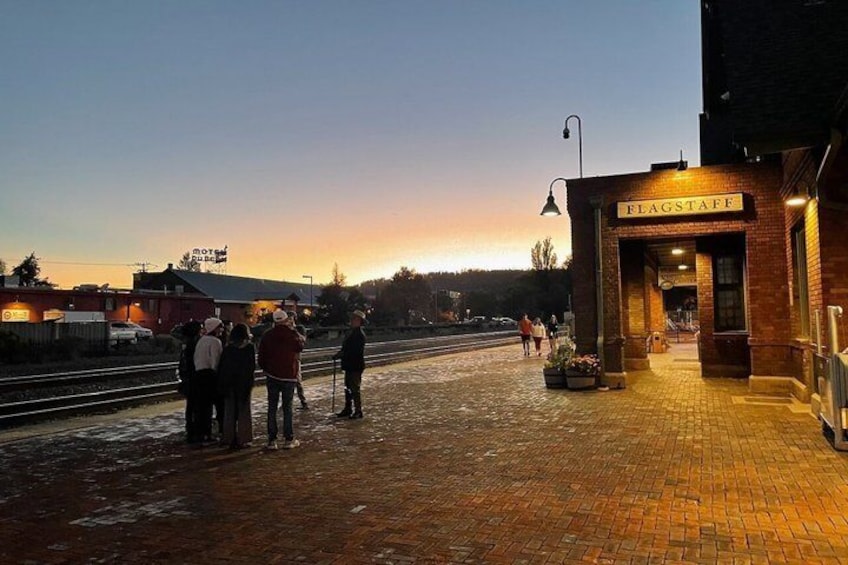 A train station platform at dusk with people waiting, a brick building labeled 'FLAGSTAFF,' and a sunset in the background. 5
