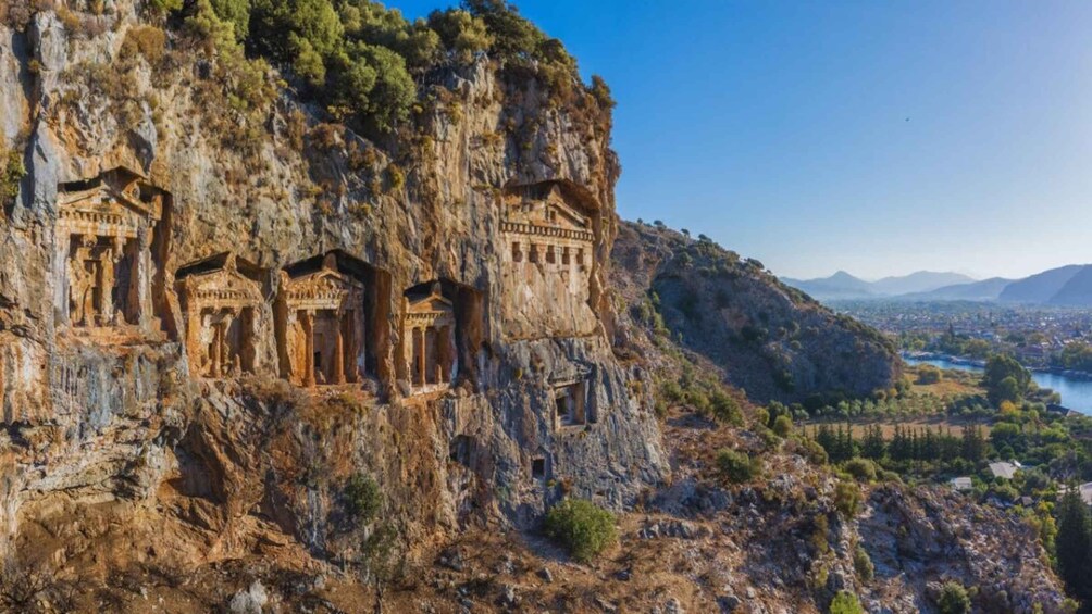 Ancient rock-cut tombs on a cliffside with a river and town in the background. 1