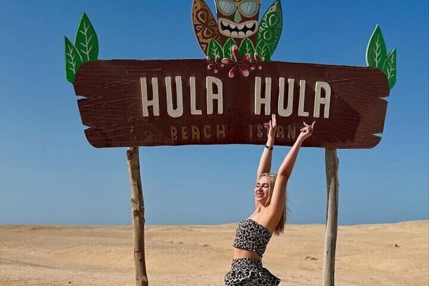 Un cartello di legno con su scritto 'HULA HULA BEACH ISLAND', con una donna che indossa una maglietta a stampa leopardo e che sta posando di fronte. 1