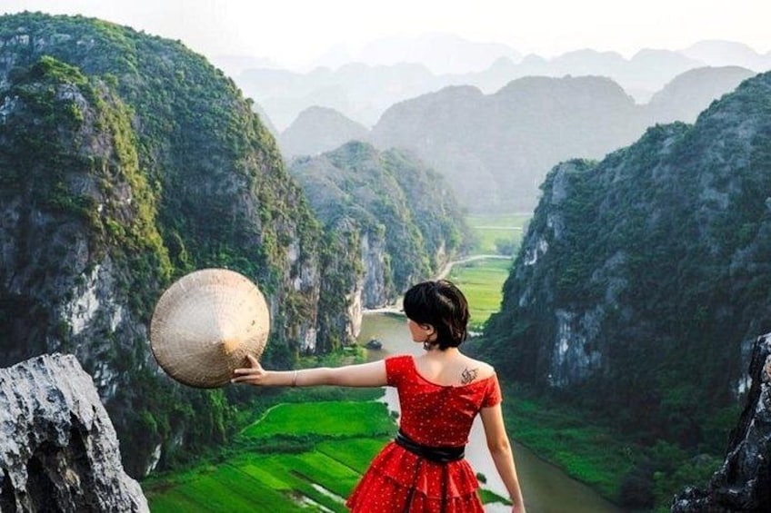 Eine Frau in einem roten Kleid hält einen großen Korb auf einer Klippe mit Blick auf ein grünes Tal. 4