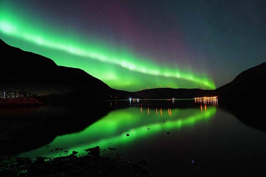 The Northern Lights over a calm lake with reflections, illuminated buildings in the background, and a mountainous landscape. 5