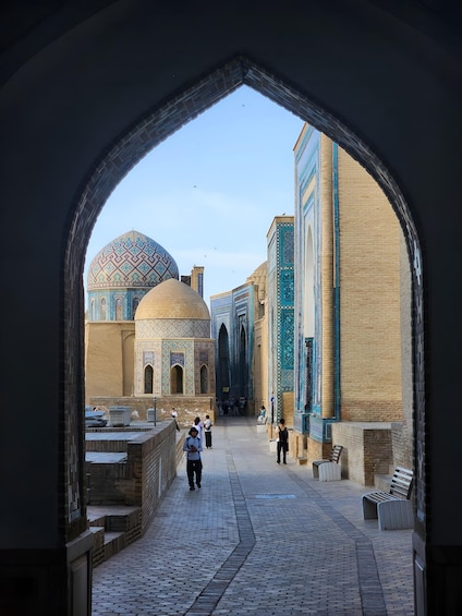 An archway leading to a courtyard with a large dome and minarets, surrounded by brick buildings with intricate tile work. 3