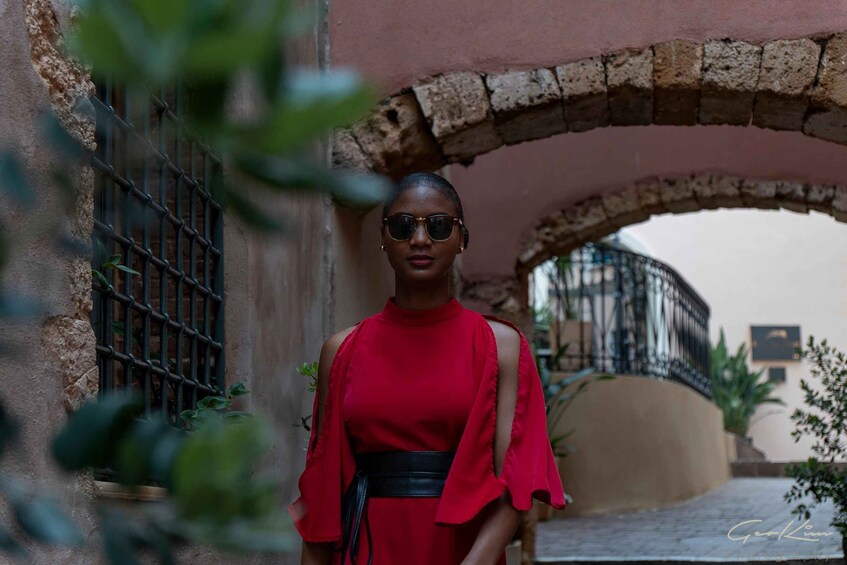 A woman in a red dress standing under a stone archway with a metal gate to the left. 2
