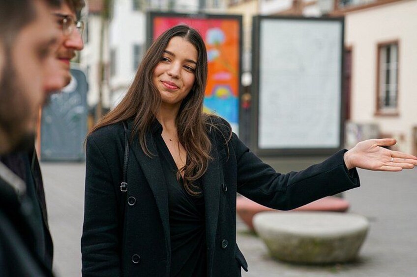Une personne fait un geste avec la main, elle porte un manteau noir, et se trouve dans un environnement urbain extérieur, avec des bâtiments et une fresque colorée en arrière-plan. 5