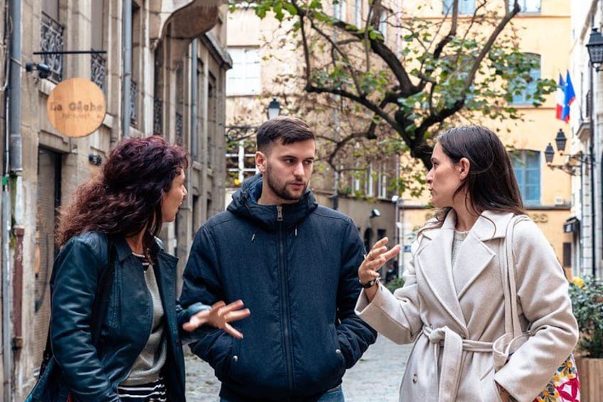 Trois personnes sont en train de discuter dans une rue de la ville, avec des bâtiments et un arbre en arrière-plan. 3