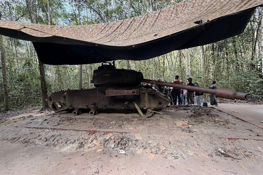 Een verroeste tank onder een zeil in een bebost gebied, met mensen in de buurt. 2