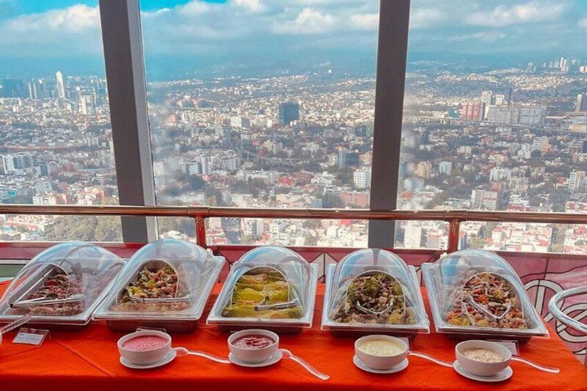 A table with food containers and bowls, a cityscape view through large windows, and a red tablecloth. 1