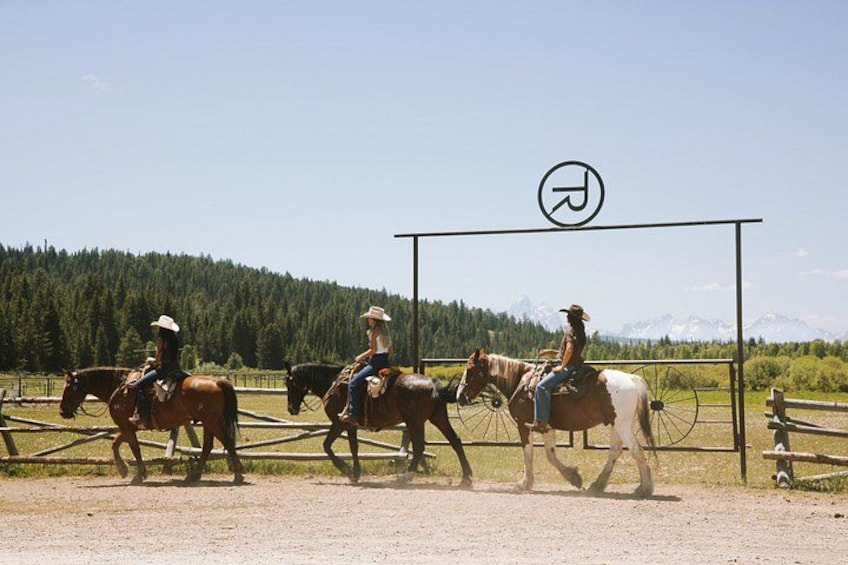 Tres personas montando a caballo por un sendero de tierra, con un bosque y montañas al fondo. 1