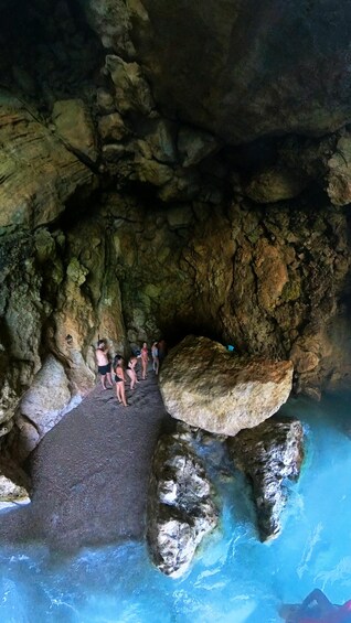 A group of people standing on a rocky ledge above a clear blue pool in a cave. 4