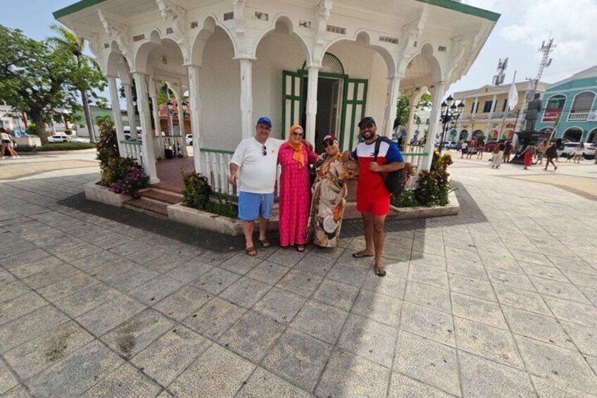 A gazebo with a white roof and green door, surrounded by a paved plaza with a statue in the background. 2
