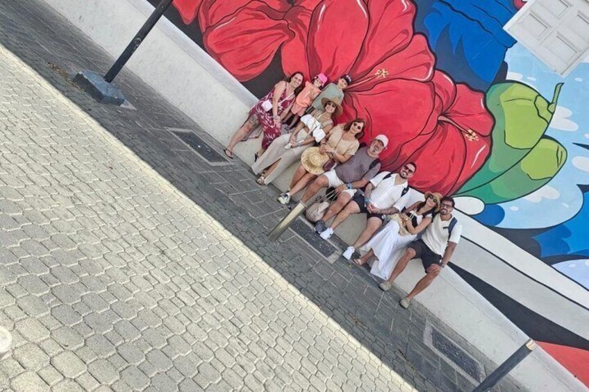 A group of people sitting on a sidewalk next to a mural with a large red flower. 5