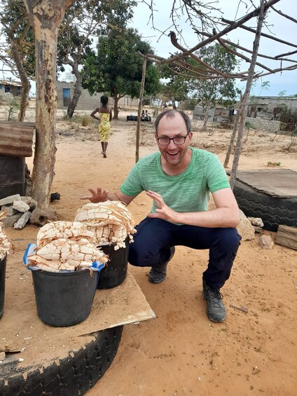 A man squatting next to two buckets filled with dried fish, with a tree and a person walking in the background. 5