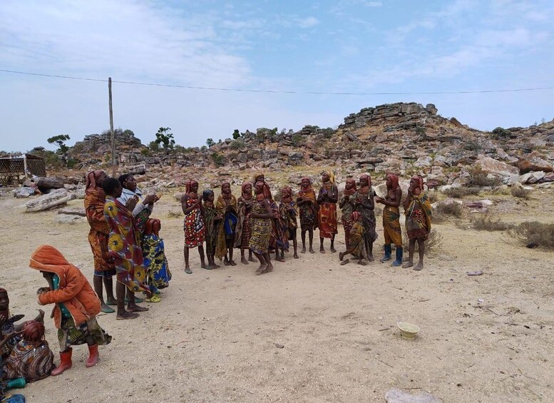 A group of people in colorful traditional attire standing in a desert-like environment with rocky terrain and sparse vegetation. 1