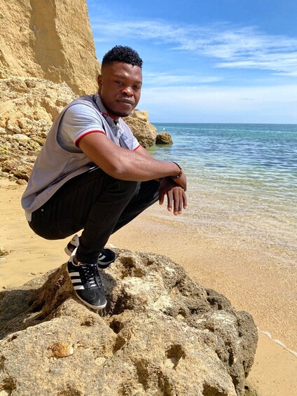 A person crouching on a rock by a sandy beach with clear water and a rocky cliff in the background. 2