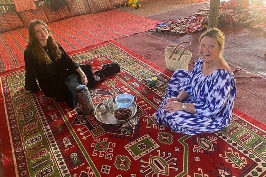 Two people sitting on a patterned rug with a tray of food and a woven basket. 4