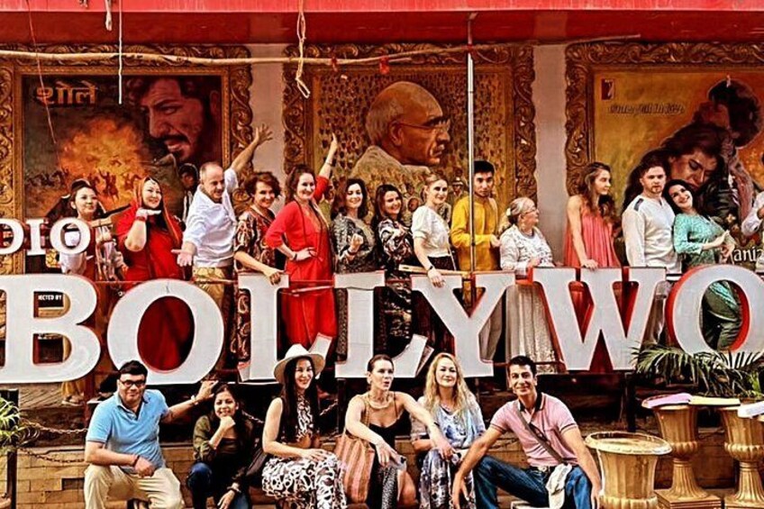 A group of people in front of a large sign with the word 'BOLLYWOOD' and a backdrop with a mural. 1