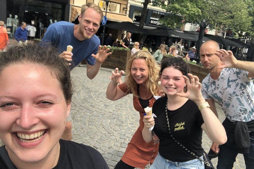 Un groupe de personnes se trouve dans un espace extérieur, sur un pavé de pierres, avec une fontaine et des devantures de magasins en arrière-plan. 2