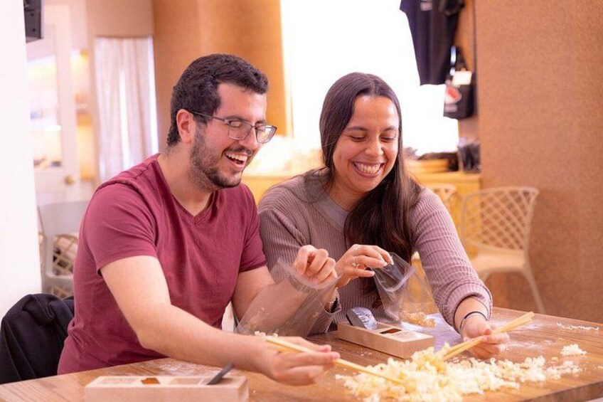 Twee personen bereiden eten, één persoon rasp kaas en de ander snijdt pasta, in een keuken met een houten tafel en stoelen. 3