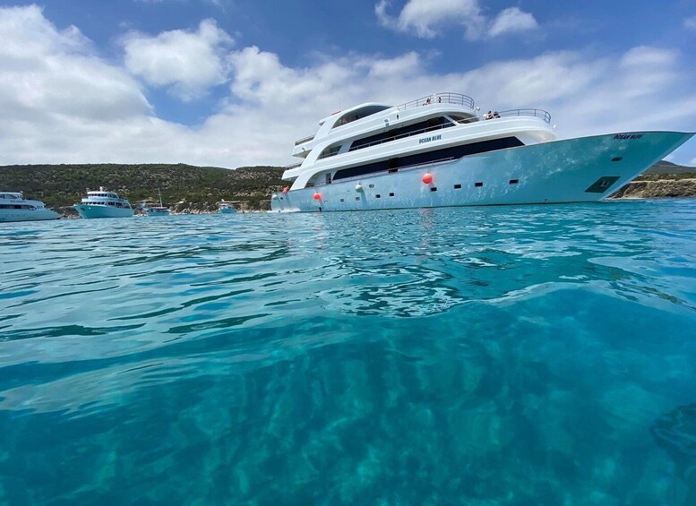 A large yacht named 'Ocean Blue' anchored in clear turquoise waters with a mountainous backdrop. 2