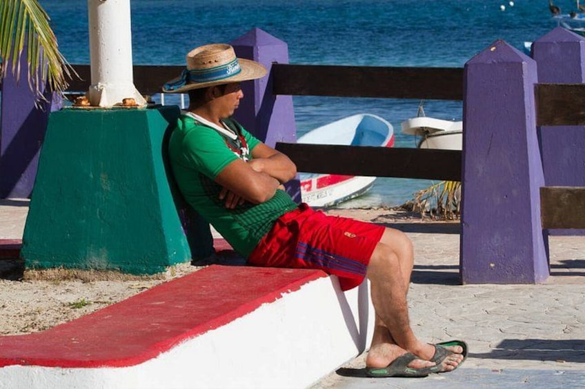 A person sitting on a bench by the sea, wearing a green shirt, red shorts, and a straw hat. 4