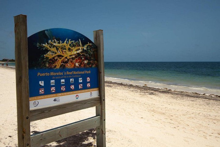 A beach sign for Puerto Morelos' Reef National Park with a clear view of the ocean and sandy beach. 2