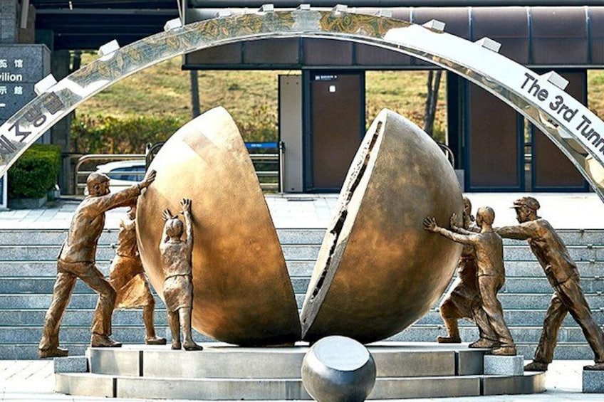 Eine Skulptur zeigt Personen, die eine große Kugel schieben, mit einem Schild, auf dem 'The 3rd Tunnel' steht. 2