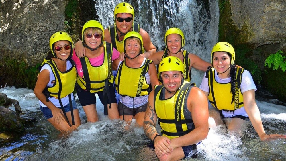 A group of people wearing yellow helmets and life vests, standing in front of a waterfall. 4
