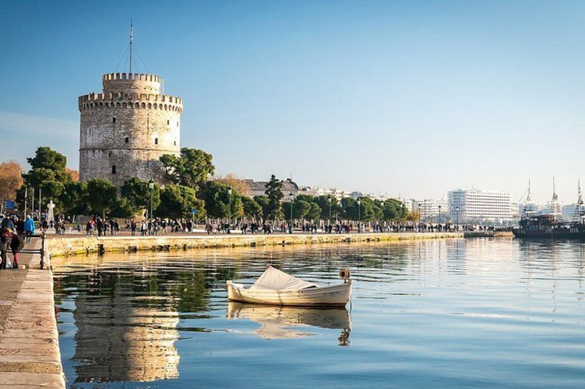A historic tower by a calm waterfront, a small boat anchored nearby, and a bustling promenade with people. 1