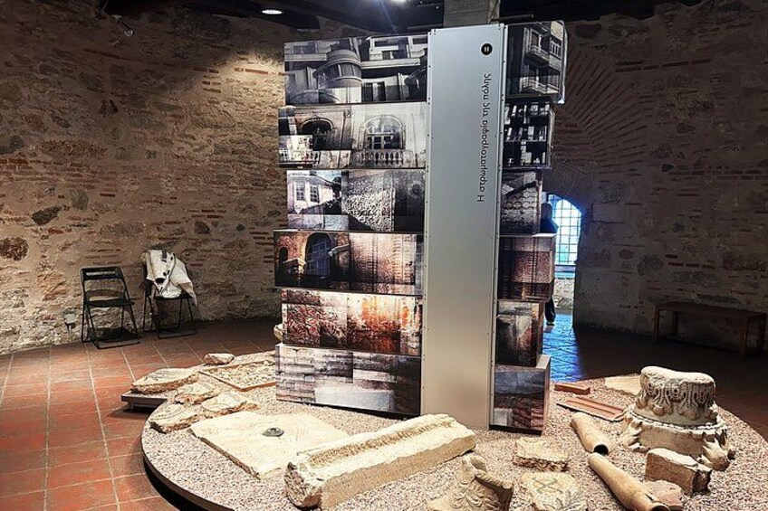 A museum exhibit with a central display of photographs and artifacts, surrounded by stone structures and a tiled floor. 4
