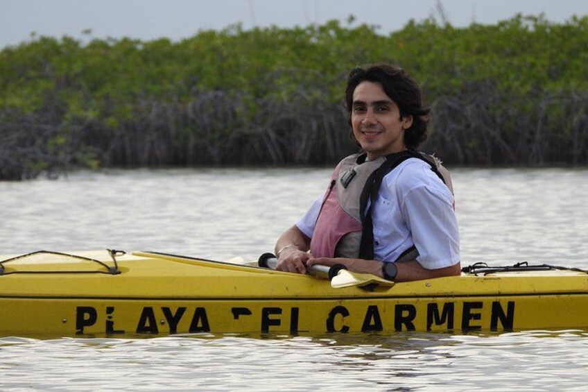 A person in a yellow kayak with 'PIAYA EF CARMEN' written on it, wearing a life jacket, on calm water with mangrove trees in the background. 5