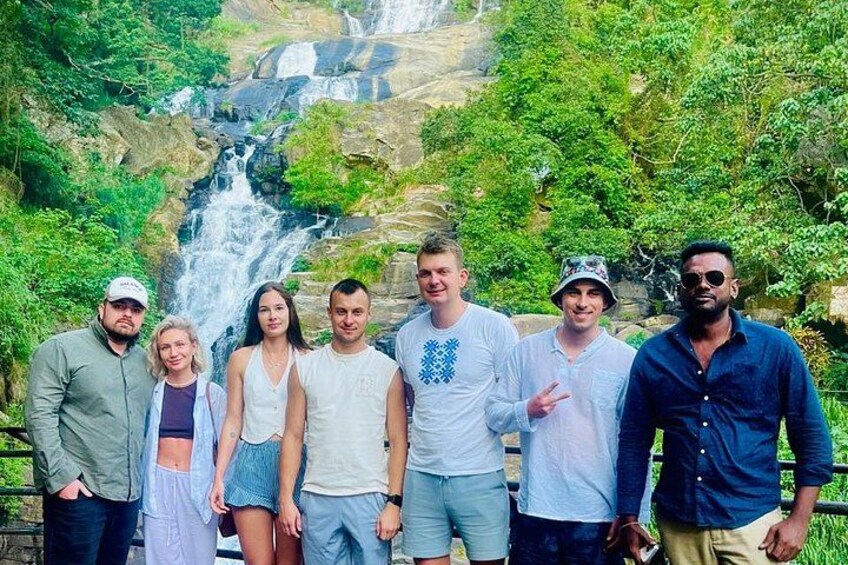 A group of people standing in front of a waterfall with lush greenery in the background. 1