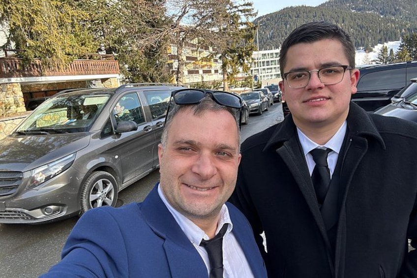 Twee personen in zakelijke kleding, een parkeerplaats en een bergachtig landschap op de achtergrond. 4