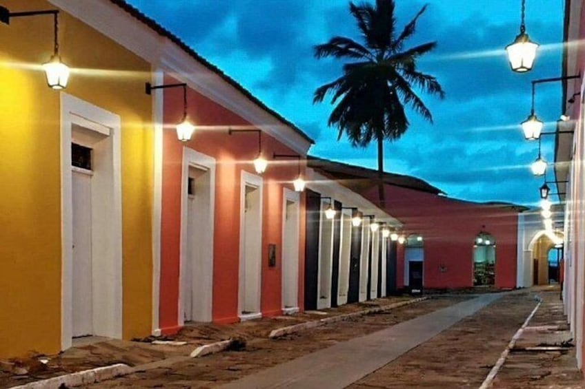 A row of brightly colored doors with hanging lights, a palm tree, and a dirt path. 3