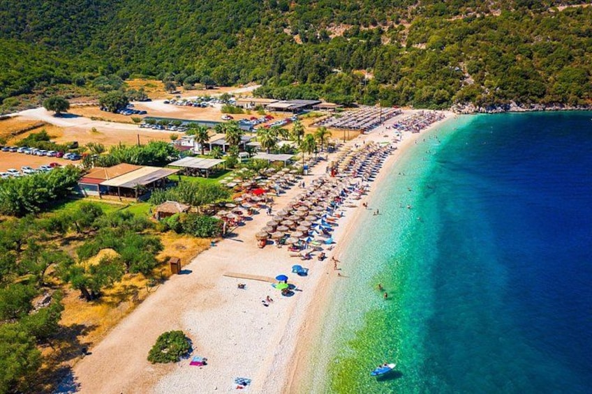 Ein Strand mit einer langen Reihe von Sonnenschirmen und Menschen, ein klares blaues Meer und Gebäude im Hintergrund. 2