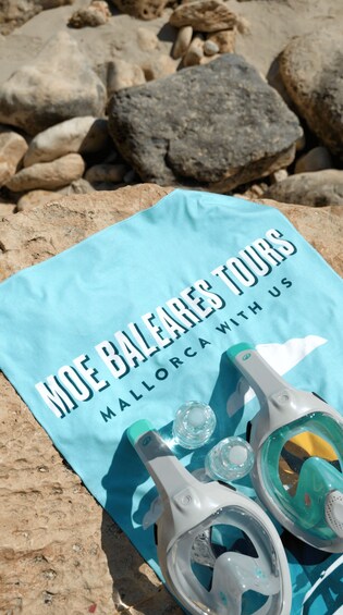 A blue towel with 'Moe Baleares Tours Mallorca' text, sunglasses, and a bottle on a rocky beach. 2
