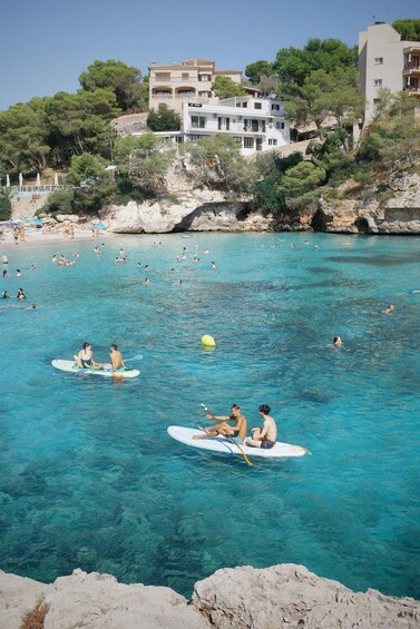 A clear blue sea with people paddling on surfboards, a rocky shoreline, and buildings in the background. 2