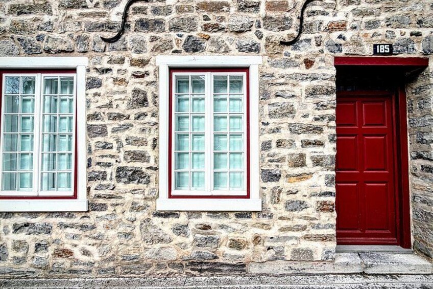 Edificio de piedra con dos ventanas y una puerta roja, visible el número 185. 1