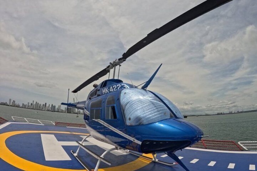 A blue helicopter with registration number HK-4223 on a helipad near a body of water with a city skyline in the background. 1