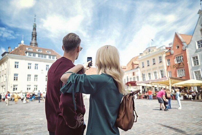 A couple taking a photo in a public square with historic buildings and a church spire in the background. 5