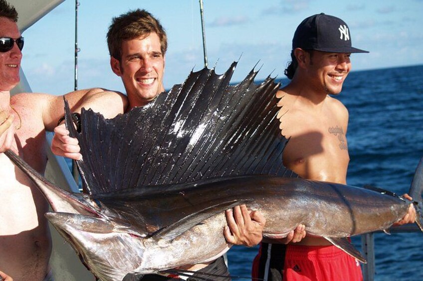 Two men holding a large fish, likely a shark, with a boat in the background. 1