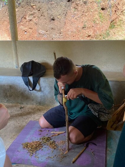 A person sitting on a mat, working with a tool and some plant material. 3