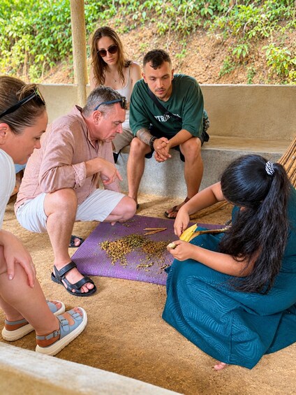 A group of people sitting on a bench outdoors, with one person cutting a banana. 2