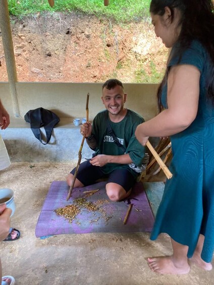 A person sitting on a mat with a stick, another person standing beside them, and scattered seeds on the ground. 5