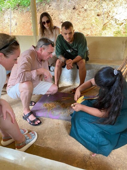 A group of people sitting on a bench outdoors, engaged in an activity with a purple cloth and some objects on the ground. 4