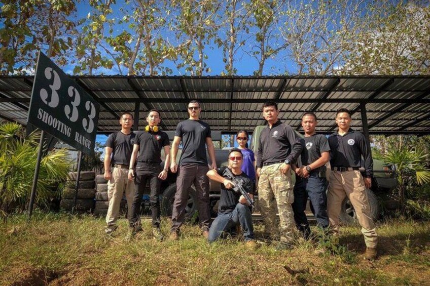 Un groupe de personnes se tient dans une zone herbeuse, devant un panneau indiquant « 333 ZONE DE TIR », et on aperçoit une structure couverte à l’arrière-plan. 2